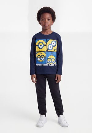 Garçon aux cheveux bouclés porte un t-shirt à manches longues bleu marine avec quatre visages de Minions et un texte, pantalon noir et baskets blanches, debout sur fond blanc.