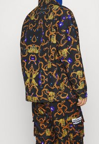 Svart jacka med ett intrikat mönster av guld- och blåkedjor, med leopardmotiv. Kombinerad med matchande byxor och en Adidas-logotyp.