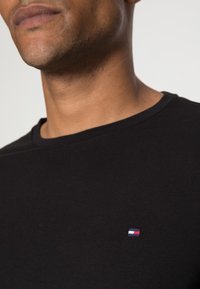 Camiseta de algodón negra con cuello redondo y un pequeño logotipo bordado en rojo, blanco y azul en el lado izquierdo del pecho.