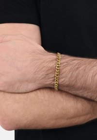 Złota bransoletka na nadgarstku, składająca się z przeplatających się ogniw o błyszczącym wykończeniu, noszona na czarnej koszuli. Gładka tekstura i wypolerowana powierzchnia.