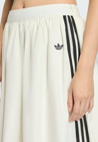 Bijela plisirana suknja s crnim Adidas prugama i logom. Ima glatku pojasnicu i sjajnu teksturu; blago se širi prema rubu.