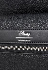 Juodas tekstūruotas odos krepšys su sidabriniu užtrauktuku, įspauduotais Disney ir Karl Lagerfeld prekės ženklais bei lygiais siuvimo elementais.
