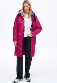 Dlouhý prošívaný kabát v jasně růžové barvě s kapucí, s vzorovanou podšívkou. Kombinovaný s tmavými džíny a šedými sportovními botami.