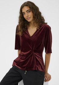 Burgundska sametna majica z V-izrezom, kratkimi rokavi in twist detajem spredaj. Gladka tekstura in sproščen kroj. Kombinirana z črnimi hlačami.