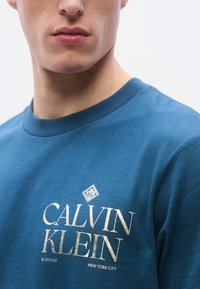 Μπλε βαμβακερό μπλουζάκι με στρογγυλή λαιμόκοψη, κοντά μανίκια και χρυσό λογότυπο με την επιγραφή «CALVIN KLEIN» και «NEW YORK CITY.»