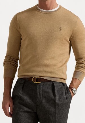 Homme portant un pull col rond couleur chameau avec un petit logo brodé, un sous-vêtement blanc, un pantalon plissé gris foncé, une ceinture marron et une montre-bracelet.