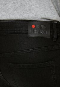 Jeans de mezclilla negra con un parche de cuero etiquetado "DENIM PROJECT" con un punto rojo. Incluye detalles de costura en el bolsillo trasero.