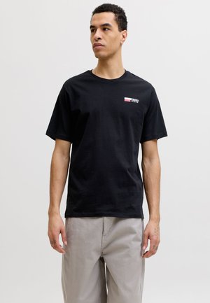 Homme debout portant un t-shirt noir Jack & Jones et un pantalon gris clair, regardant légèrement vers la gauche sur un fond uni.