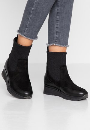 Person trägt schwarze Keilabsatz-Stiefeletten, kombiniert mit grauen, kurz geschnittenen Jeans mit fransigen Säumen und steht auf einem glänzenden Boden.