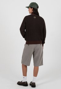 Brun teksturert genser med ribbet kant, kombinert med grå shorts og mørke slip-on sko. Grønn lue fullfører antrekket, sett bakfra.