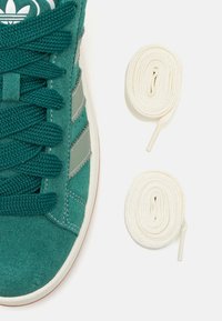 Grüne Wildledersneaker mit strukturiertem Material, ausgestattet mit weißen Schnürsenkeln und auffälligen silbernen Streifenakzenten. Zwei zusätzliche gerollte cremefarbene Schnürsenkel sind enthalten.