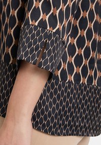 Granatowa bluzka z geometrycznym wzorem, przedstawiająca diamenty w odcieniach kremu i brązu. Krótkie rękawy ze szczelinami, gładki materiał i luźny krój.