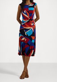 Mujer vistiendo un vestido midi sin mangas con un patrón geométrico abstracto multicolor y detalle retorcido en la cintura, combinado con sandalias marrones.