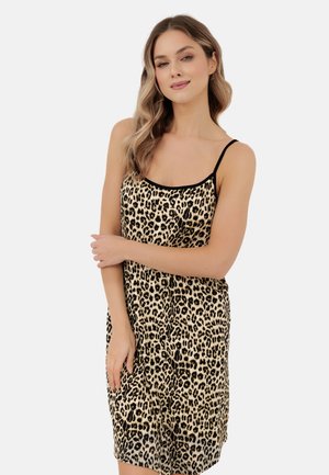 Kobieta o falowanych blond włosach, ubrana w sukienkę bez rękawów w panterkę z cienkimi czarnymi ramiączkami, stojąca ze skrzyżowanymi ramionami na białym tle.