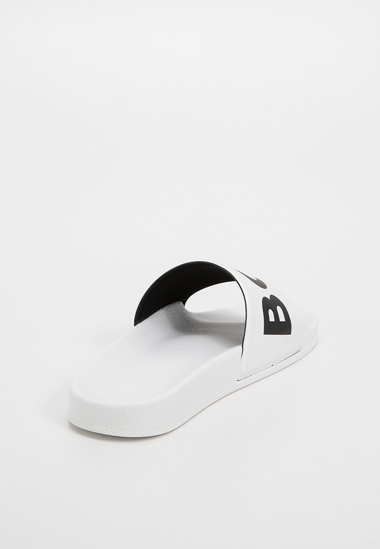 Savates blanches à glissière avec une sangle noire, affichant une grande lettre "B" noire sur le côté. Texture lisse avec une semelle plate en caoutchouc durable.