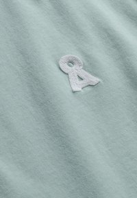 White embroidered "A" logo on light green textured fabric, showing fabric weave and soft folds.