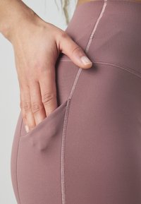 Leggings mauve a vita alta realizzati in un tessuto morbido ed elastico, dotati di una tasca laterale e cuciture a contrasto.