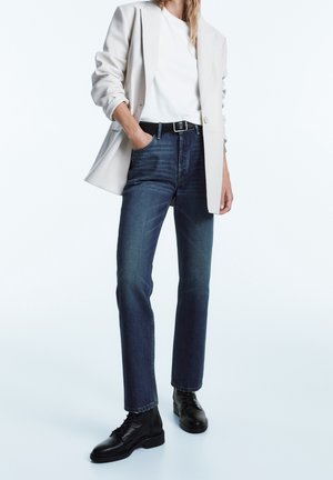 Person iført lys beige blazer, hvid skjorte, mørkeblå jeans med sort bælte og sorte snørestøvler, stående med den ene hånd i lommen.