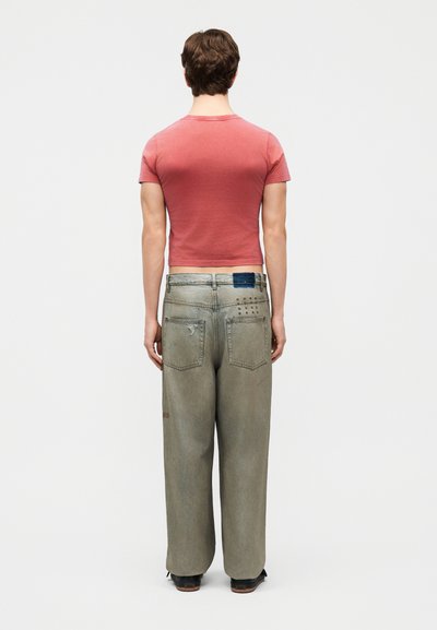 Muž stojaci chrbtom, oblečený v priliehavom vyblednutom červenom tričku a voľných opotrebovaných svetlosivých džínsoch s zadnými vreckami a tmavými topánkami.