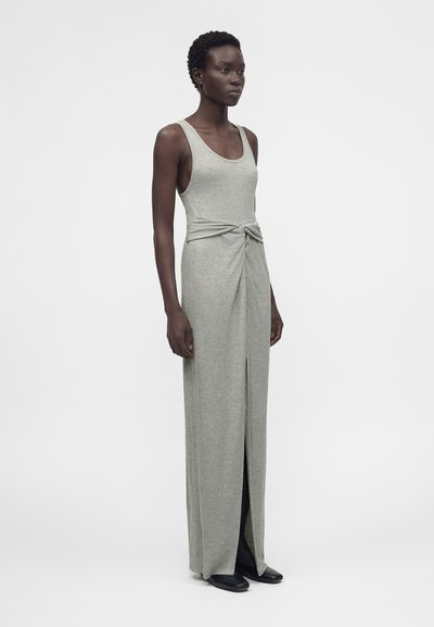 Γυναίκα που στέκεται, φοράει ασύμμετρο αμάνικο maxi φόρεμα ανοιχτού γκρι χρώματος με κόμπο μπροστά και σκίσιμο, συνδυασμένο με μαύρα παπούτσια, σε απλό φόντο.