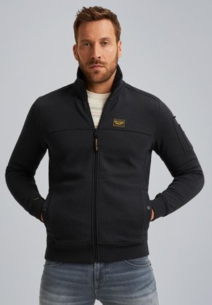 Black zip-up jacket with a textured panel at the chest, smooth sleeves, side pockets, and a small logo patch above the left pocket.