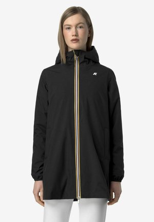 Kobieta w czarnej, kapturowej pelerynie przeciwdeszczowej z pionowym zamkiem błyskawicznym w paski i elastycznymi mankietami, stojąca na tle jednolitego, jasnego tła.