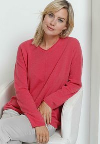 Rosa V-Ausschnitt Pullover mit langen Ärmeln, aus einem weichen, strukturierten Stoff. Kombiniert mit hellen Hosen, sitzt in einem weißen Stuhl.