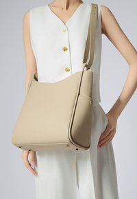 Borsa a spalla in pelle beige con una texture liscia, forma curva, tracolla regolabile e dettagli in metallo dorato.