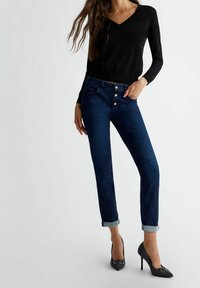 Dunkelblaue Jeans mit einem Knopfverschluss, leicht hochgeschlagenen Säumen, kombiniert mit einem schwarzen V-Ausschnitt-Pullover und gemusterten High Heels.
