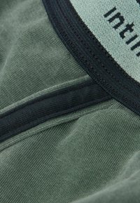 Grünes Baumwollkleidungsstück mit weicher Textur, ausgestattet mit einem schwarzen elastischen Bund und kontrastierenden Nähdetails.