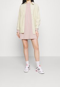 Ljust beige skjorta med knappar över en rosa kort klänning, i kombination med vita sneakers med grå detaljer och röda inslag; vita ankelsockor.