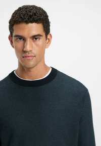 Dark blue knit sweater with a textured finish, featuring a ribbed crew neckline. Smooth white inner collar is visible beneath the sweater.