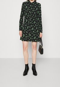 Czarna sukienka w kwiaty z zielonymi wzorami, z guzikami z przodu, długimi rękawami i ściągniętą talią, w połączeniu z czarnymi botkami za kostkę i torebką.