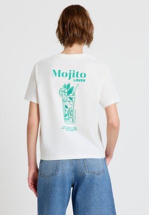 Persona con capelli corti che indossa una maglietta bianca con un'illustrazione di un bicchiere di mojito turchese e il testo "Mojito Lover" sulla schiena.