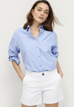Kvinde med skulderlangt brunt hår iført en lyseblå skjorte med knapper, ærmerne rullet op, og hvide shorts, stående med hænderne i lommerne.