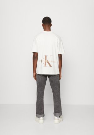 Weißes Baumwoll-T-Shirt mit großem beigem Calvin Klein Jeans-Logo auf der Rückseite, kombiniert mit grauen Hosen und weißen Sneakern.