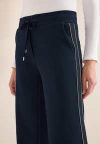 Granatowe spodnie dresowe wykonane z tkaniny o fakturze, z bocznymi kieszeniami, sznurkiem w talii i białym obszyciem wzdłuż nóg.