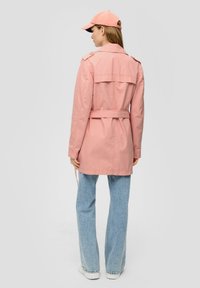 Lichtroze trenchcoat met een ceintuur in de taille, schouderepauletten en een achterflapdetail, gecombineerd met lichtblauwe wide-leg jeans en witte sneakers.