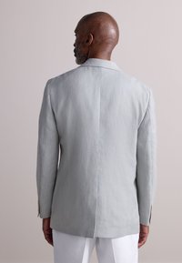 Blazer in lino grigio chiaro con finitura testurizzata, spacco singolo sul retro e due bottoni sul polsino. In primo piano su uno sfondo neutro.