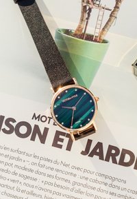 Okrągły zegarek z zieloną paskową tarczą i złotymi akcentami, z czarnym teksturowanym paskiem i kryształowymi znacznikami na tarczy.