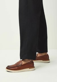 Mocassins en cuir marron avec couture décorative, semelle rembourrée et semelle beige, portés avec un pantalon noir court.