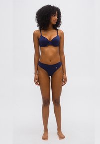 Femme aux cheveux bouclés portant un soutien-gorge bleu marine assorti à sa culotte, debout pieds nus contre un fond clair uni, regardant sur le côté.