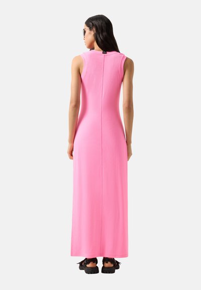 Kvinde med langt mørkt hår iført solbriller og en ærmeløs, lys pink maxikjole, stående med ryggen mod beskueren.