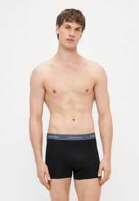 Musta puuvillainen bokserialushousu, jossa on harmaa elastinen vyötärönauha, jossa on "Calvin Klein" -logo, sileä pinta ja istuva muotoilu.