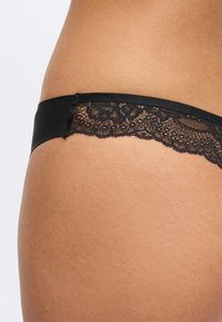 Sous-vêtements noirs à dentelle portés sur la hanche, montrant une peau lisse et un motif floral détaillé en dentelle sur le panneau latéral.