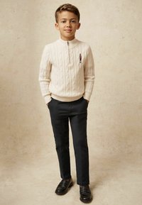 Krém színű, cable-knit pulóver, cipzáras gallérral, sötét, slim-fit nadrággal és fekete loaferekkel párosítva. Figyelemre méltó hímzett részlet a pulóveren.