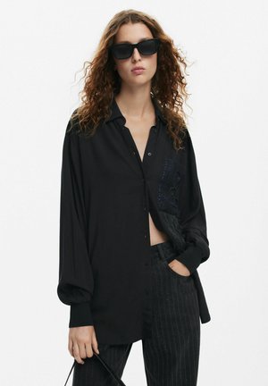 Camicia nera con bottoni, caratterizzata da un dettaglio tasca testurizzato, con vestibilità comoda e maniche lunghe. Abbinata a pantaloni a righe scure.