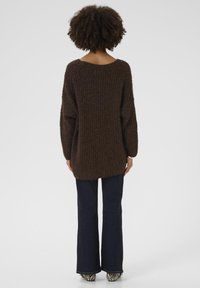 Brown, oversized knitted sweater with a ribbed texture. Features long sleeves and a wide neckline, paired with dark flared jeans and striped shoes.