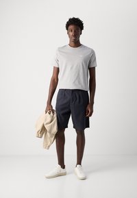 Tricou gri din bumbac, pantaloni scurți bleumarin, jachetă bej și adidași albi purtați de o figură masculină în picioare, pe un fundal deschis.