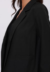 Blazer noir en tissu texturé, avec une poche poitrine unique, des revers crantés et une coupe ajustée.